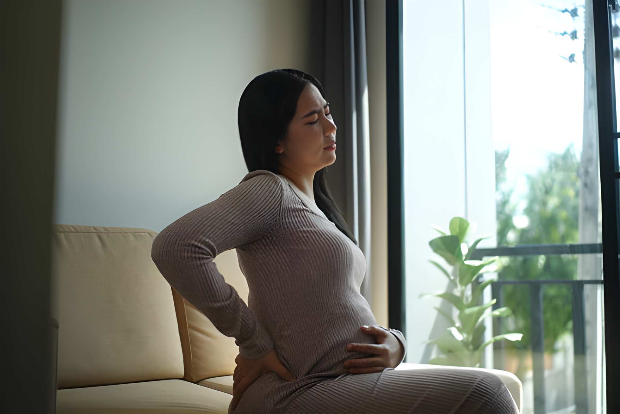Pregnant woman sitting on a sofa holding her lower back due to lower back pain during pregnancy.
